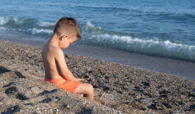 Niño triste sentado solo en la playa de arena cerca del mar.