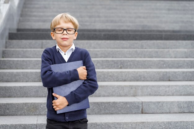 Un niño con uniforme azul y gafas está parado en los escalones con un cuaderno azul