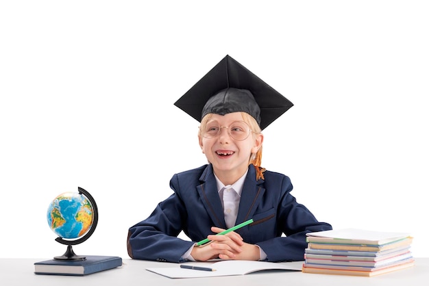 Niño en uniforme escolar y sombrero de estudiantes se sienta en el escritorio y se ríe sobre fondo blanco Educación en universidad universitaria en el extranjero