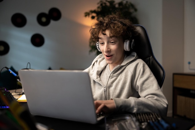 Un niño usa una computadora portátil en el escritorio. El niño con el cabello rizado usa audífonos. Está escuchando música. Jugando juegos de computadora. Navegando por Internet. Charlando con amigos.