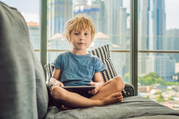 Niño usa una tableta en casa en el sofá en el fondo de una ventana con rascacielos. Los niños modernos de la megalópolis usan un concepto de tableta