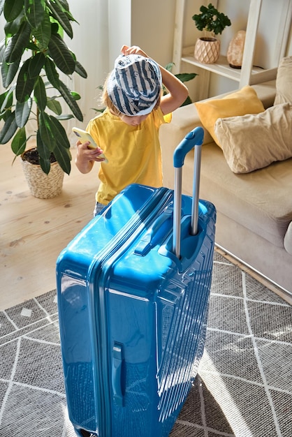 Niño usando un teléfono móvil comprando boletos en línea o esperando vacaciones en casa junto a una maleta empacada