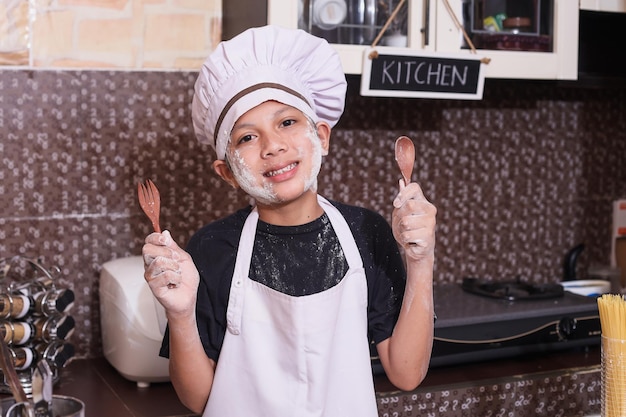 Niño usando uniforme de chef sosteniendo cuchara y tenedor de madera, haciendo masa en la cocina, harina en las mejillas.