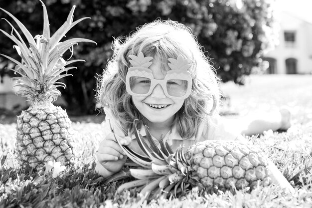 Niño en vasos divertidos con fruta de piña tendido sobre el césped en el fondo de la naturaleza de verano