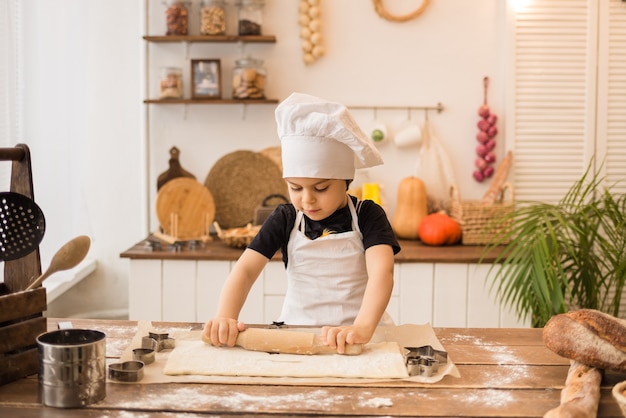 Niño vestido como un chef extiende la masa con un rodillo de madera en la mesa de la cocina