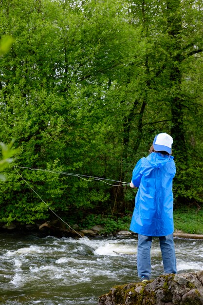 Niño, vestido con impermeable azul y gorra, pescando en el río de montaña. Vista desde atrás