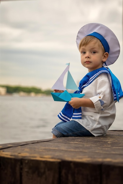 Un niño vestido de marinero se sienta en la orilla con un barco de papel
