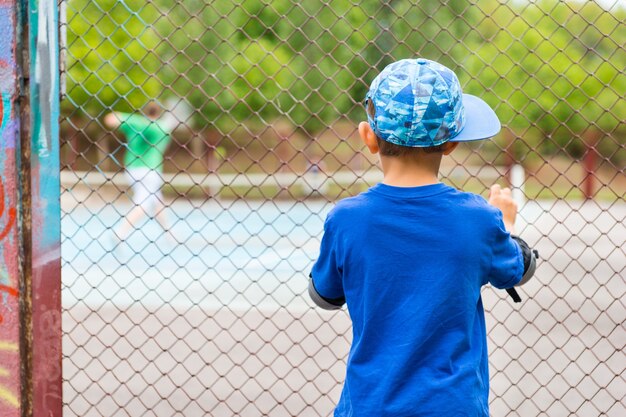 Niño viendo un partido de tenis de pie de espaldas a la cámara mirando a través de una valla de malla de alambre a los jugadores