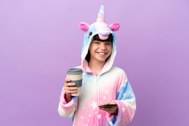 Niño vistiendo un pijama de unicornio aislado sobre fondo púrpura con café para llevar y un móvil