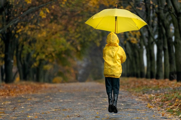 Niño vuela o salta con un gran paraguas amarillo Niño camina bajo la lluvia en un parque de otoño Vista posterior