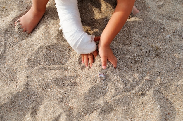 Niño con yeso en la mano jugando en la arena de la playa junto al mar.