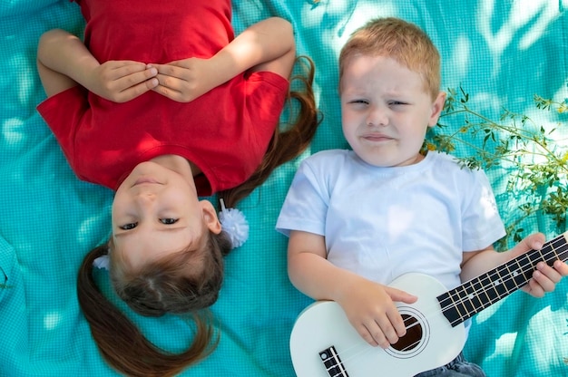 Los niños se acuestan en el césped del parque en verano y tocan el ukelele Momentos felices guitarra primer plano bebé Un niño alegre y encantador disfruta de un saludable juego de verano al aire libre