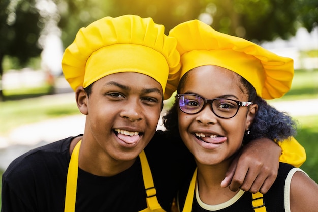 Los niños africanos cocinan con sombrero de chef y uniformes amarillos haciendo muecas y mostrándose la lengua unos a otros Adolescente africano y niña negra se divierten y cocinan