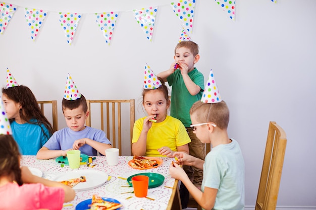 Niños alegres comiendo pizza en una mesa grande los niños usan sombreros de cumpleaños divirtiéndose juntos sabrosa pizza para la fiesta de los niños