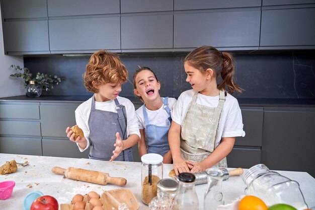 Niños alegres con delantales amasando masa para galletas en una mesa desordenada con varios ingredientes en una cocina moderna y luminosa en casa