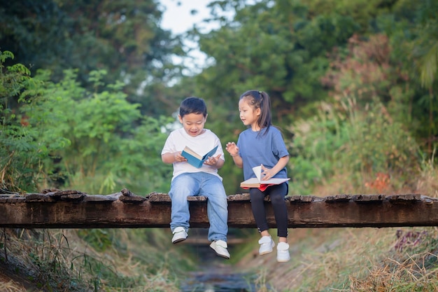 Niños alegres sentados en un puente de madera Niños asiáticos jugando en el jardín niño y niña leyendo libros