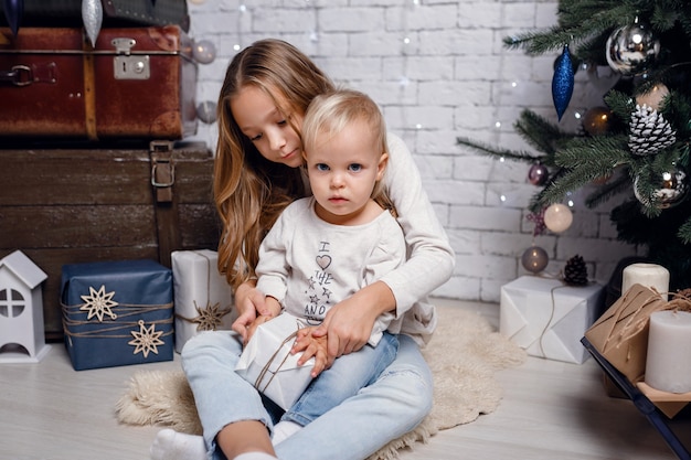 Niños bajo el árbol de Navidad con cajas de regalo. Decoraciones de año nuevo. estilo rústico, decoraciones de madera oscura. Interior de año nuevo. Hermanas.