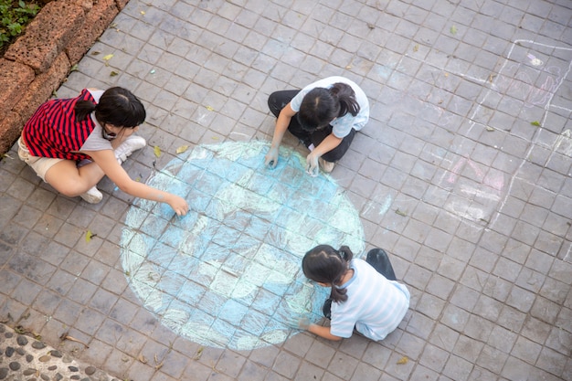 Los niños asiáticos juegan al aire libre. La niña dibuja un globo terráqueo con un mapa del mundo con tiza de colores sobre el pavimento, asfalto. Tierra, concierto del día de la paz.