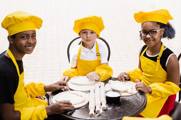 Niños cocineros multiétnicos con sombrero de chef amarillo y delantal cocinando masa para panadería Niños negros africanos y caucásicos cocinando y divirtiéndose juntos