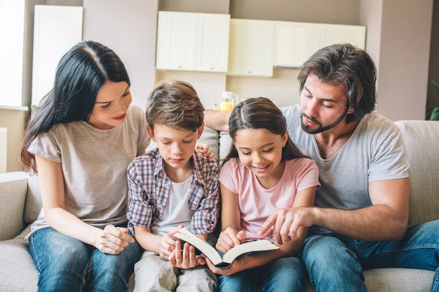 Foto niños concentrados están sentados en el sofá con sus padres y leyendo un libro.