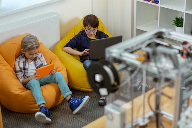 Niños concentrados sentados cómodamente en bolsas de sillas brillantes y trabajando con dispositivos