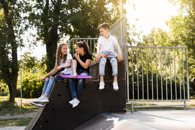 Los niños deportistas con patinetas y pennyboards están sentados y charlando entre ellos en una rampa deportiva en el patio de recreo Comunicación infantil y amistad Deporte estilo de vida extremo