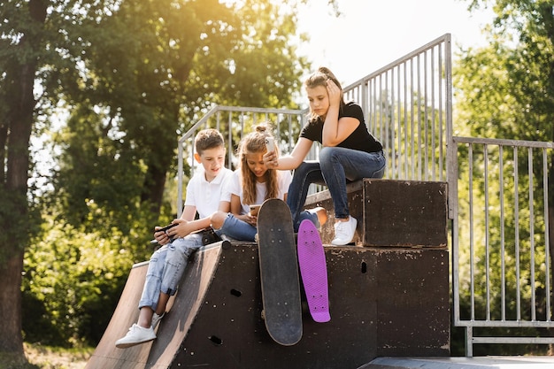 Los niños deportivos adictos al teléfono con monopatín y tableros de centavos están sentados y mirando teléfonos inteligentes en la rampa deportiva en el patio de recreo