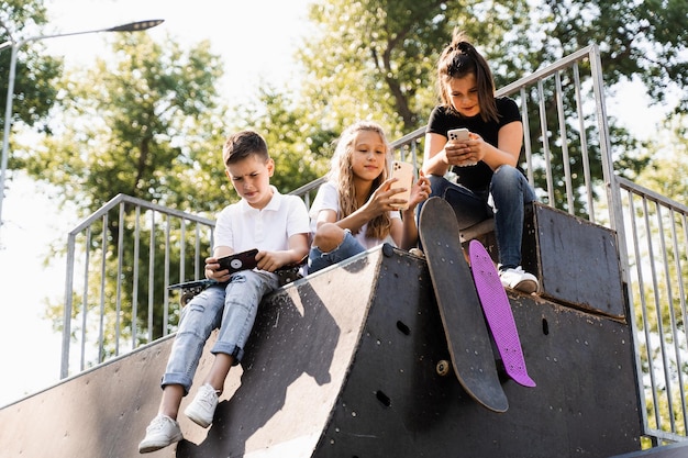 Los niños deportivos adictos al teléfono con monopatín y tableros de penny están sentados y mirando teléfonos inteligentes en la rampa deportiva en el patio de recreo