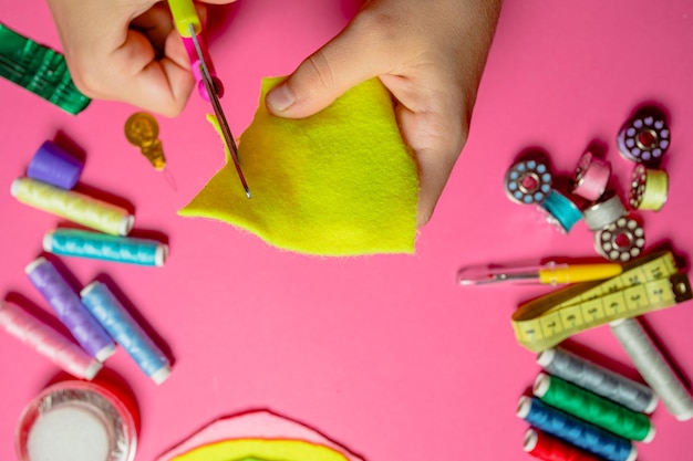 Niños diseñando y haciendo regalos para familiares y amigos Costura como pasatiempo Manos de niños cortando hilos de colores de fieltro amarillo en carretes y otros materiales de costura colocados sobre fondo rosa