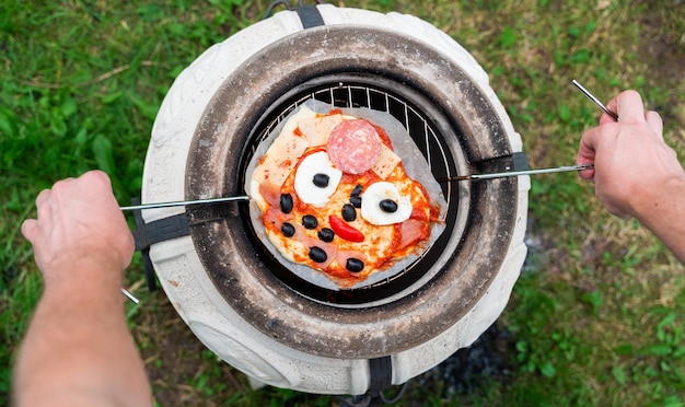 Niños divertidos pizza casera con cara ojos de queso aceitunas negras boca de tomate salsa de salchicha Cocina familiar al aire libre en horno oriental tandoor Rejilla metálica especial para barbacoa verduras Hierba verde