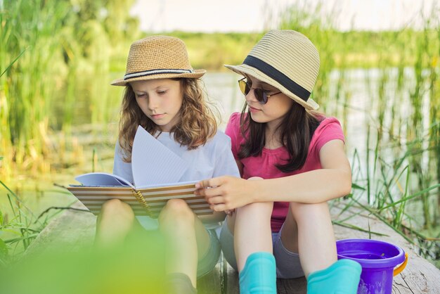 Niños dos niñas descansando jugando leyendo su cuaderno en la naturaleza. Niños sentados en el muelle del lago de madera, fondo de paisaje de agua de atardecer de verano, estilo campestre