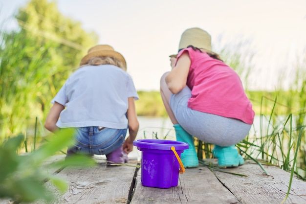 Los niños dos niñas juntos jugando con agua en el lago en el muelle de madera en cañas. Niños con balde recogiendo palos de algas y caracoles de agua. Naturaleza, ocio, amistad, concepto de vacaciones de verano