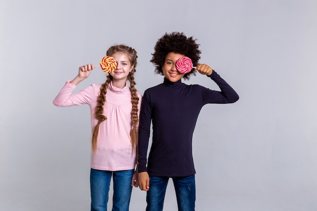 Niños con dulces. Funky niños pequeños de pie juntos y jugando con caramelos mientras está de pie sobre fondo gris