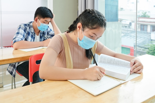 Los niños de la escuela vietnamita en máscaras médicas leyendo libros y escribiendo en cuadernos durante la lección