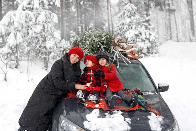 Los niños están sentados en el capó de un automóvil con un árbol de Navidad y un regalo y una madre está de pie al lado.