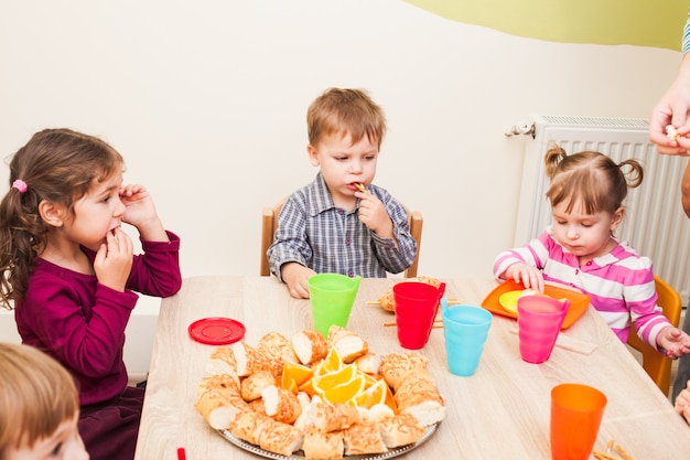 Los niños están sentados a la mesa con el almuerzo y comiendo frutas y pasteles.