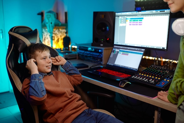 Niños en el estudio de grabación preadolescente escuchando una nueva banda sonora sentados en una mesa de escritorio con equipo de producción de sonido