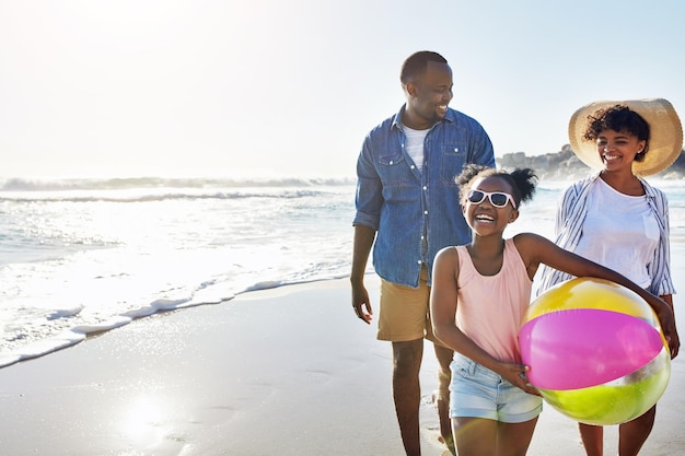 Niños de familia negros o playa con una madre, padre e hija que llevan una pelota mientras caminan sobre la arena junto al mar. Aman la naturaleza y el océano con un hombre, una mujer y una niña en la costa en verano.