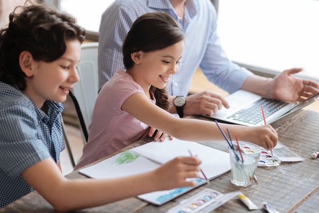 Niños felices y agradables sentados en la mesa y pintando un cuadro de acuarela juntos mientras su padre trabaja en la computadora portátil