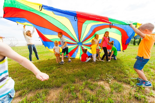 Niños felices en camisetas coloridas ondeando en paracaídas arco iris sobre sus amigos