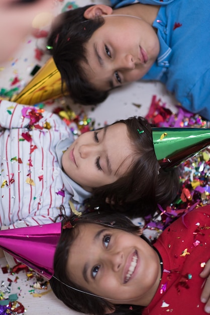 Niños felices celebrando la fiesta con confeti soplando mientras están tirados en el suelo