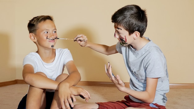 Niños felices contentos divirtiéndose y dibujando con pincel en la cara sentada en el piso