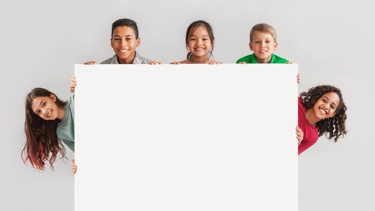 Niños felices y diversos escondidos detrás de un fondo gris de tablero blanco vacío