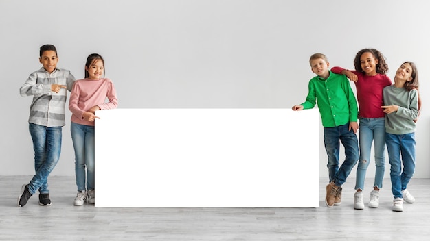 Niños felices y diversos parados cerca de una pizarra blanca sobre fondo gris