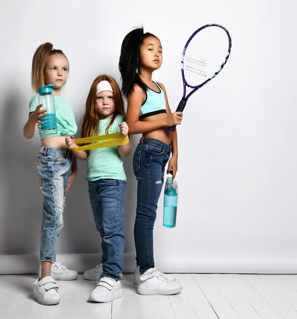 Niños felices con equipos deportivos retrato de tiro de estudio aislado sobre fondo blanco Niñas multiétnicas en jeans camisetas deportivas sostienen una botella de agua una gran raqueta de tenis una banda de fitness