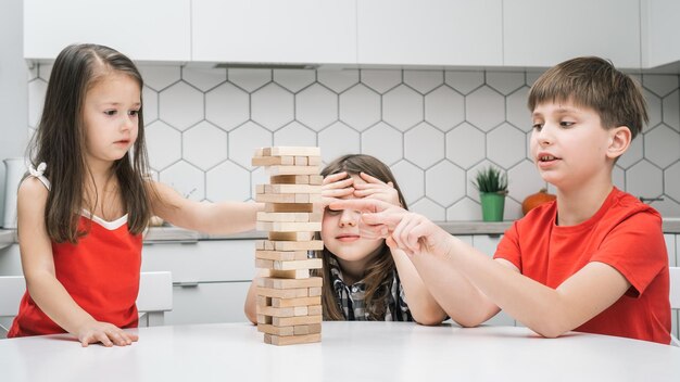 Los niños felices de la escuela juegan a la torre sentados en la mesa de la cocina Lindos niños y niñas construyen una torre con pequeños bloques de madera