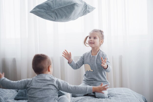 Niños felices jugando en el dormitorio blanco. Niño y niña, hermano y hermana juegan en la cama en pijama. Pijamas y ropa de cama para bebés y niños pequeños. Familia en casa.