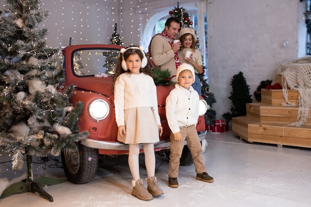 Niños felices jugando con nieve cerca del coche rojo y el árbol de Navidad, sus padres están cerca de ellos. Feliz navidad y felices fiestas