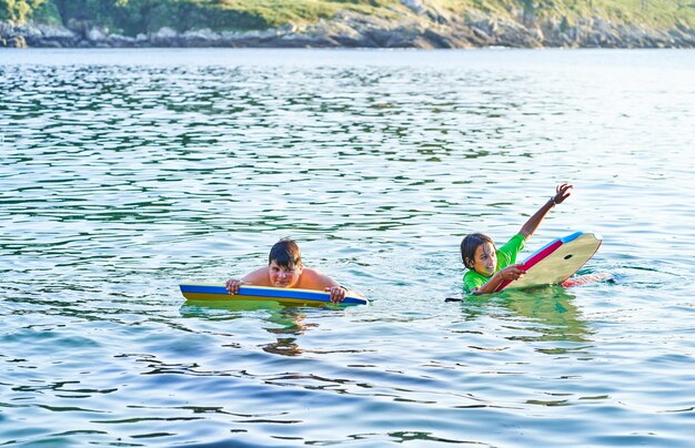 Niños felices mirando a cámara jugando en el mar con bodyboard. Niños divirtiéndose al aire libre. Concepto de vacaciones de verano y estilo de vida saludable.