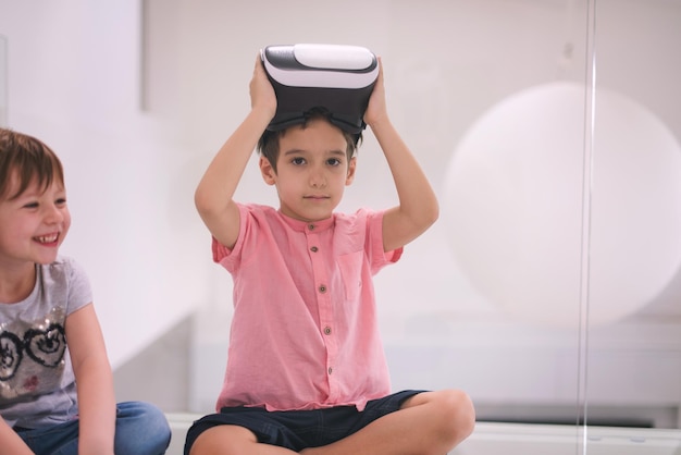 Niños felices que usan auriculares de realidad virtual y señalan con el dedo mientras se sientan en el suelo en el interior de una casa moderna y luminosa